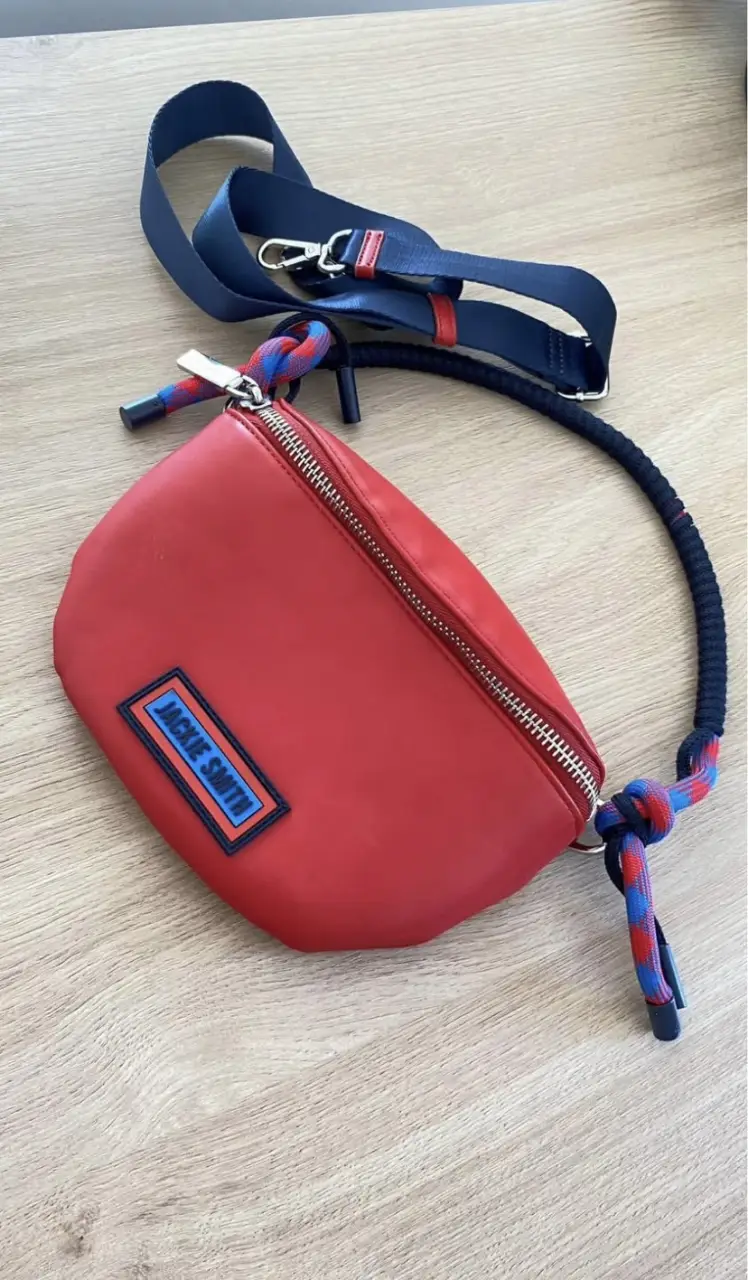 Riñonera Jackie Smith roja con detalles en azul y blanco. Cuenta con un bolsillo principal con cierre y un parche con el logo de la marca. Incluye correa ajustable y detalles en los extremos.