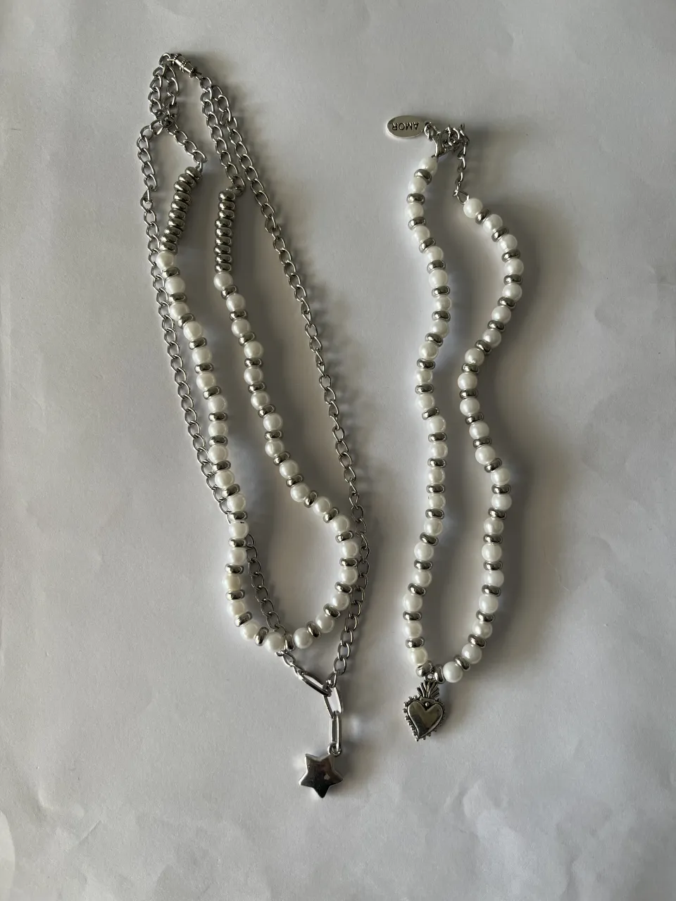 Dos collares, uno con cadena plateada y perlas blancas con un dije de estrella. El otro collar es de perlas blancas con un dije de corazón.