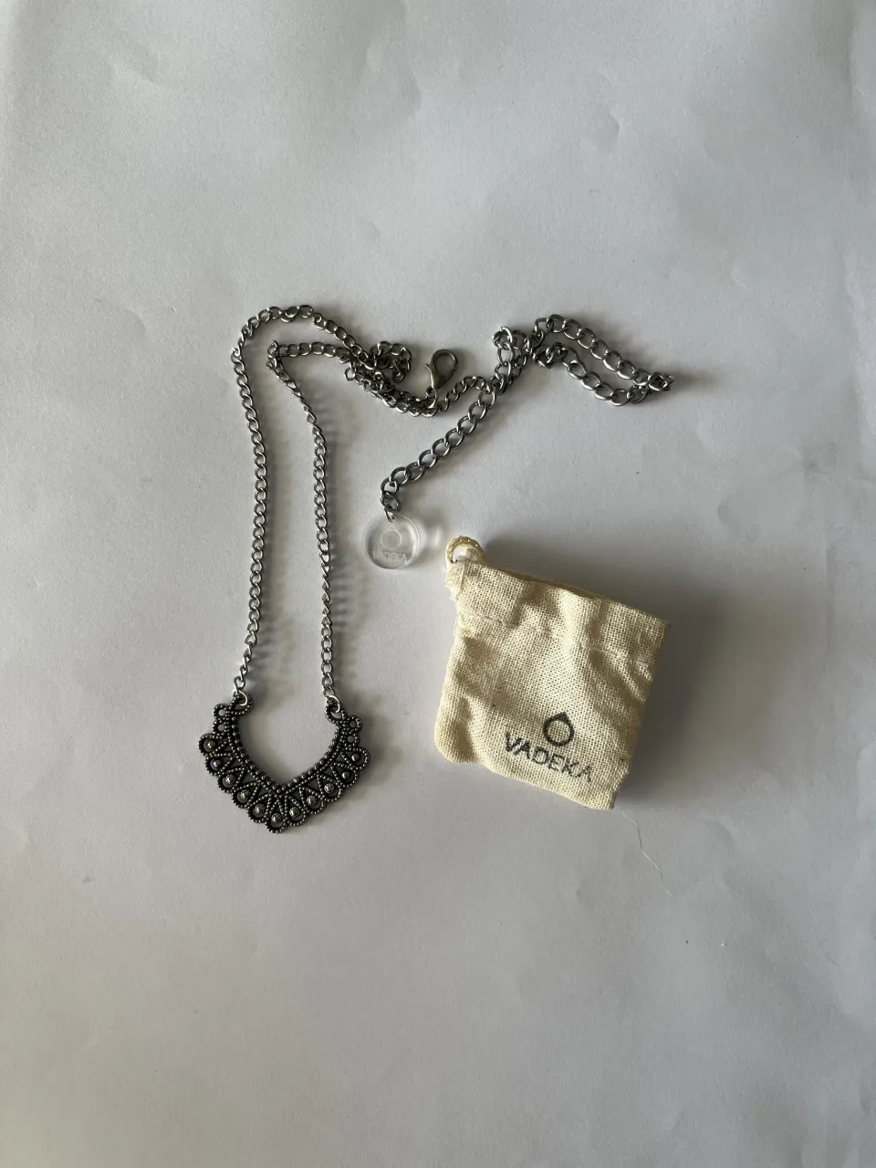 Collar plateado con dije de diseño curvo y detalles en relieve, acompañado de una bolsita de tela color crudo con logo. 