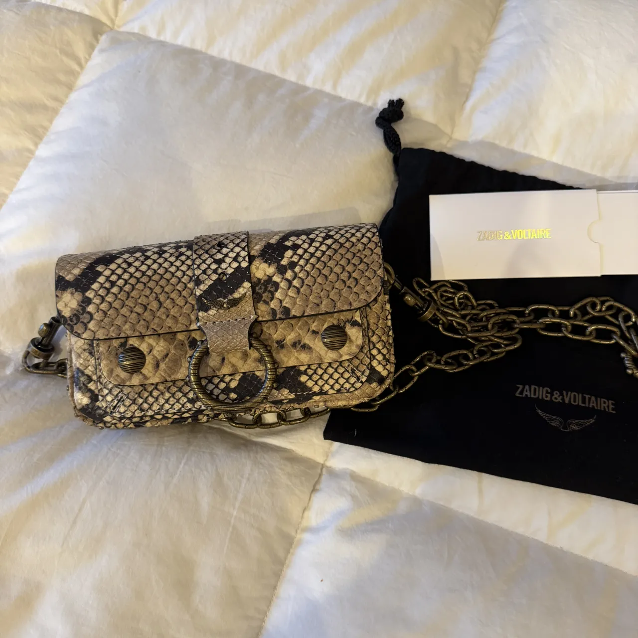 Cartera mini de cuero con estampado de serpiente, ideal para llevar lo esencial. Posee detalles metálicos y una cadena para usar cruzada o al hombro. El interior está firmado, lo que le da un toque único y personal.