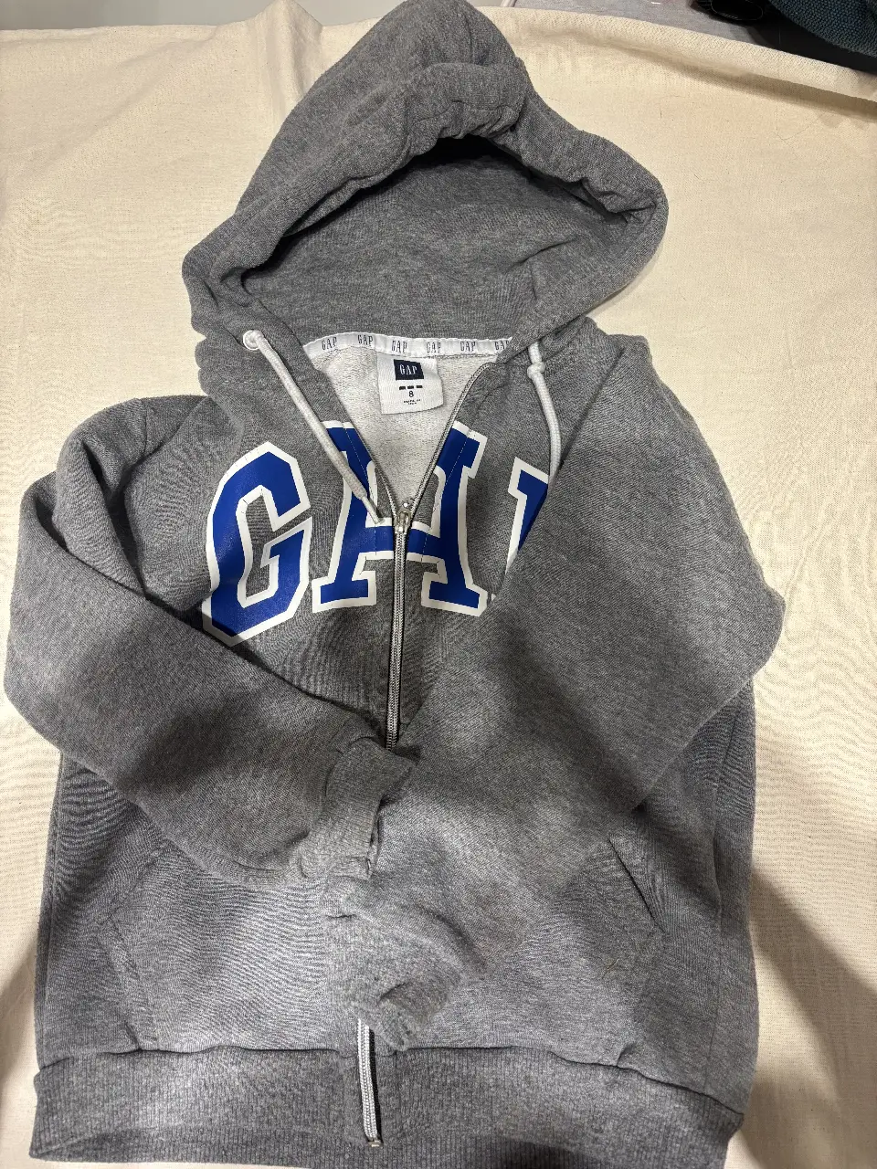 Buzo con cierre y capucha de la marca GAP, color gris con letras azules. Ideal para un look casual y cómodo.