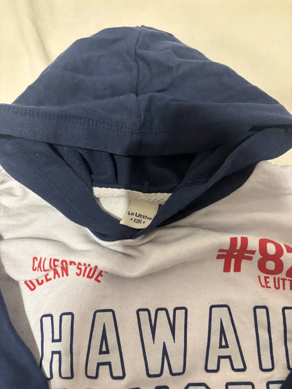 Buzo con capucha para niño, de color blanco con mangas y capucha azul marino. Estampado "Hawaii Sunset" en el frente y detalles en rojo. Ideal para un look casual y cómodo.