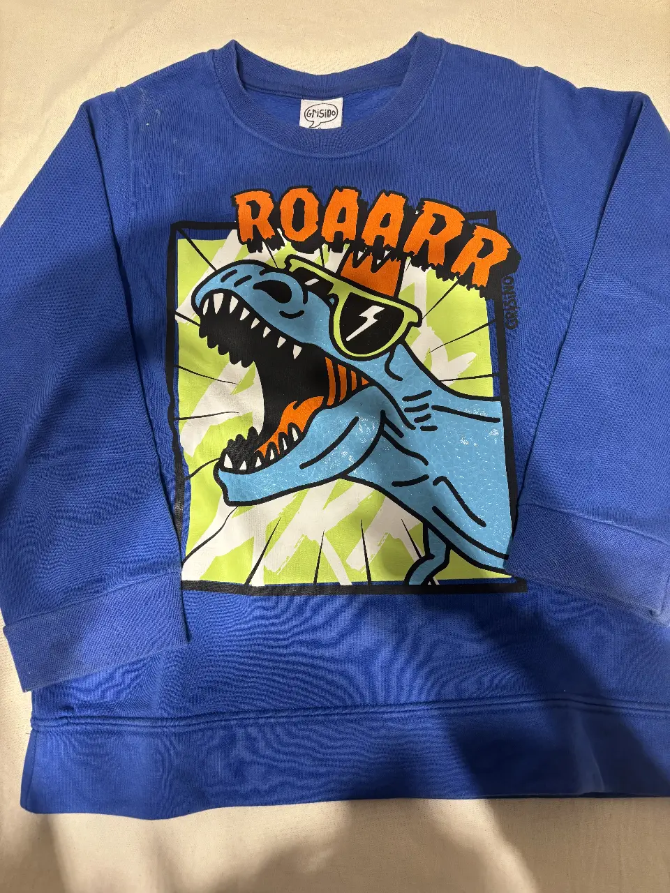 Buzo de cuello redondo para niño, de color azul con estampado de dinosaurio "ROAARR" con lentes de sol. Ideal para un look casual y divertido.