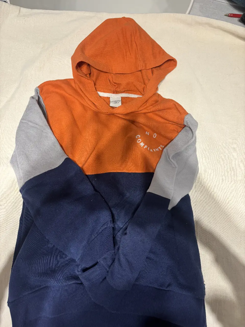 Buzo canguro para niño, con capucha y diseño en bloques de color naranja, gris y azul marino. Estampado "NO COMPLAINTS" en el frente. Ideal para un look casual y cómodo.
