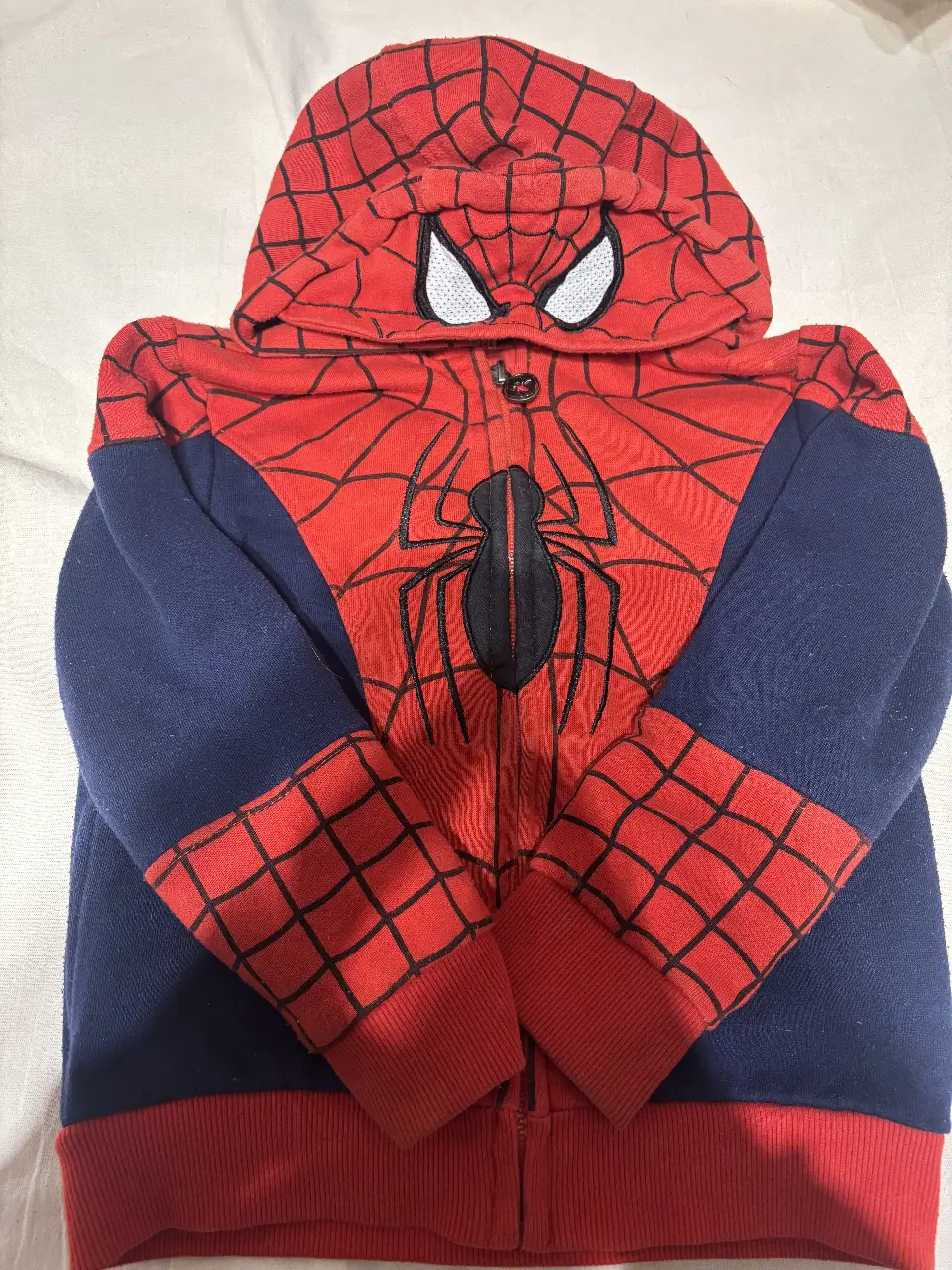 Buzo con capucha de Spiderman para niño. Diseño con cierre frontal, capucha con máscara de Spiderman y detalles de telaraña. Mangas en azul marino y cuerpo en rojo con el logo de Spiderman.