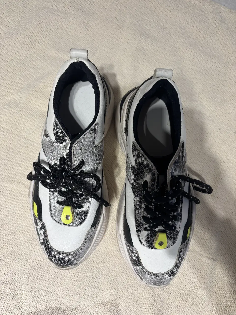 Zapatillas deportivas blancas con detalles en negro y estampado de serpiente. Cordones negros y detalles amarillos. Suela gruesa y diseño moderno.