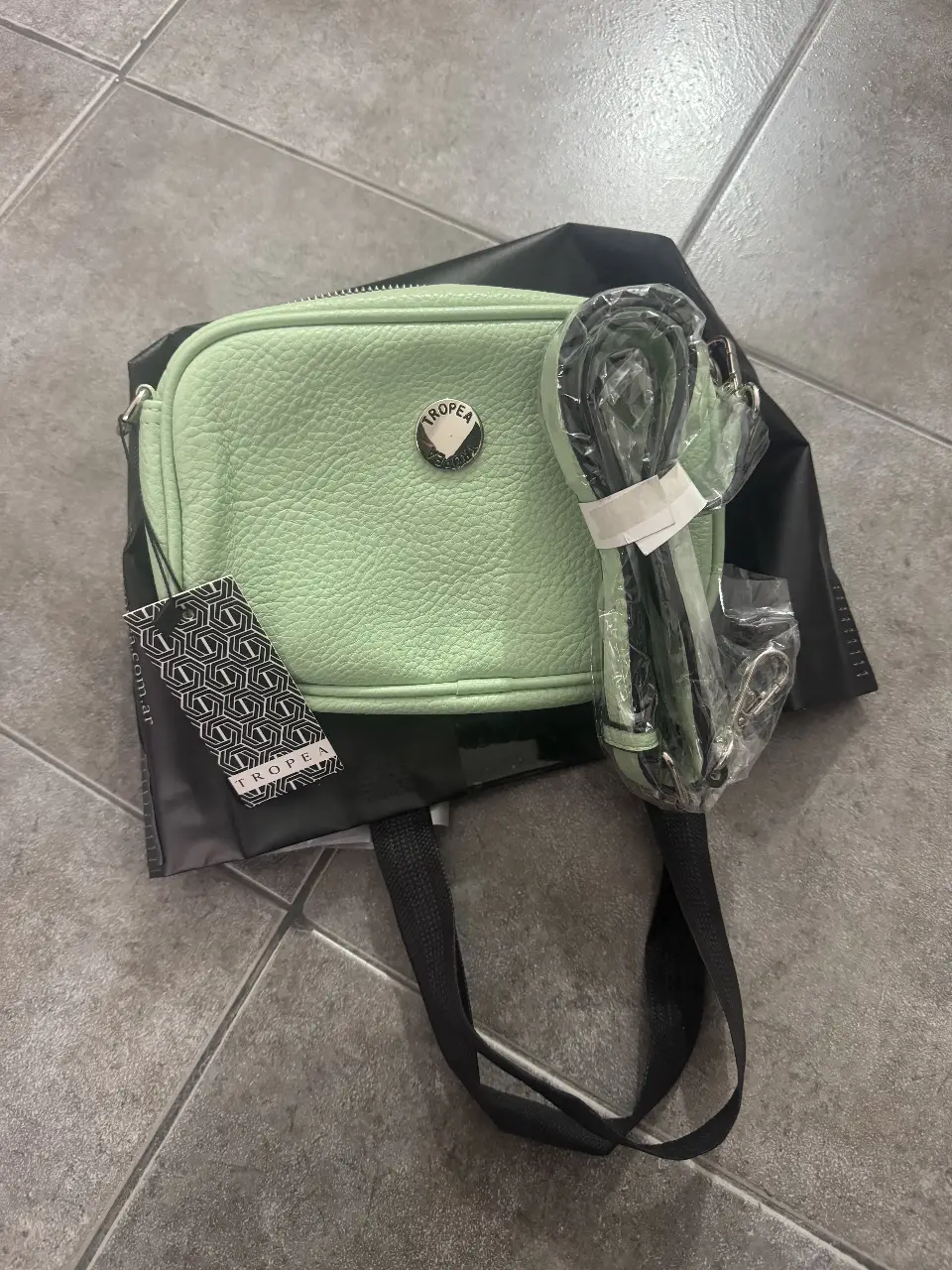 Cartera bandolera de cuero sintético color verde agua, con correa ajustable y etiqueta de la marca Tropea. Ideal para llevar tus esenciales con estilo.
