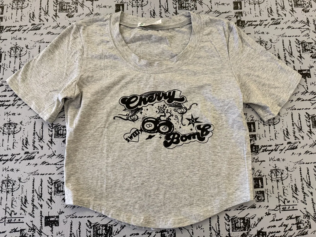 Remera gris con estampa "Cherry Bomb" y "Sweet" en la parte delantera. La estampa incluye dos bolos de pool con el número 8, un corazón, estrellas y elementos decorativos.