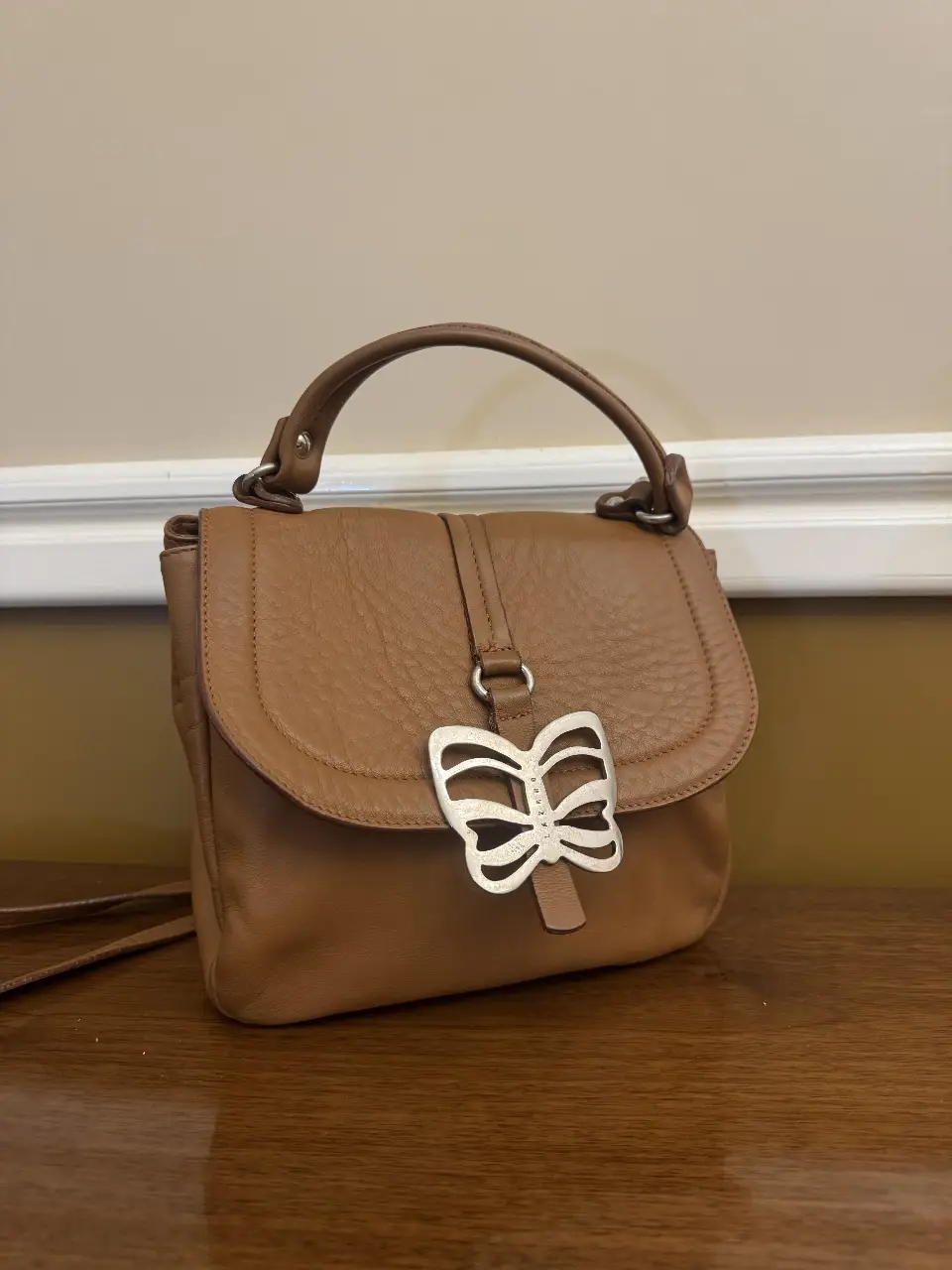 Cartera de cuero marrón con detalle de mariposa plateada. Ideal para un look casual y elegante. Cuenta con manija superior y correa para llevar al hombro.