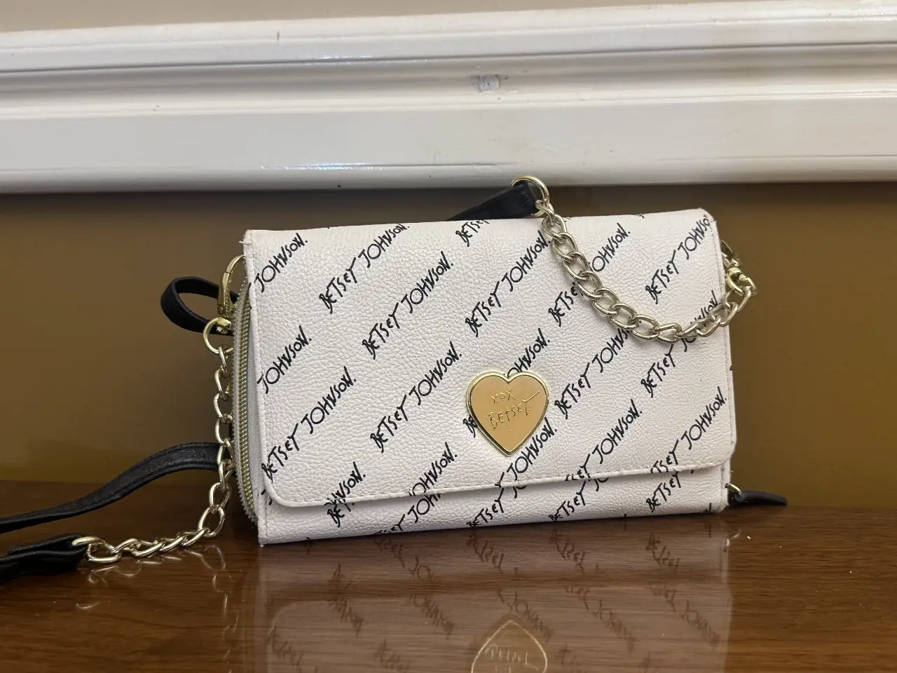 Cartera bandolera de mujer, color blanco con estampado del logo Betsey Johnson en negro. Detalles dorados, incluyendo el dije de corazón y la cadena. Ideal para un look casual y moderno.