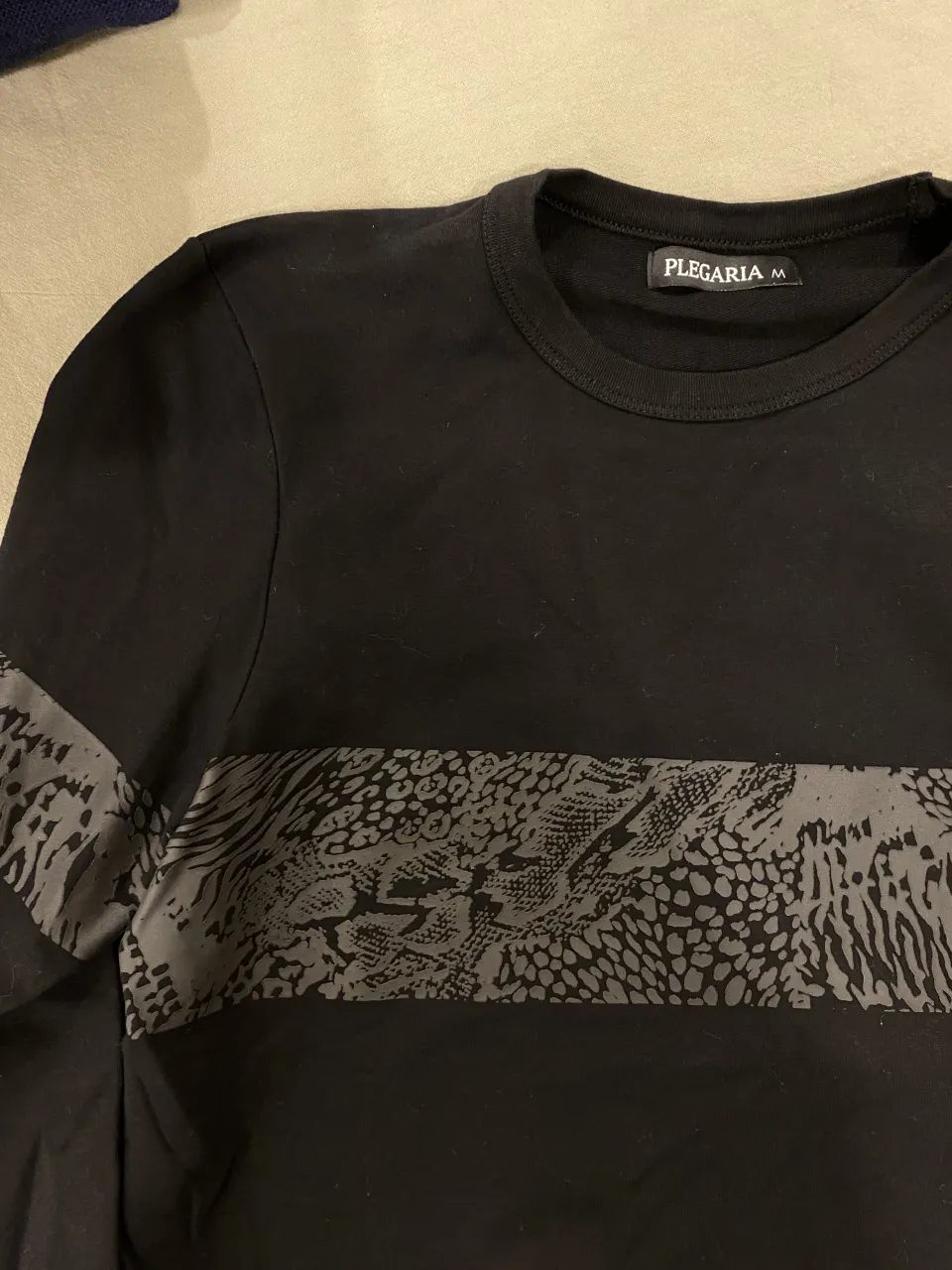 Remera negra de manga larga con una franja horizontal estampada en el frente. La estampa simula piel de serpiente y tiene detalles texturizados.