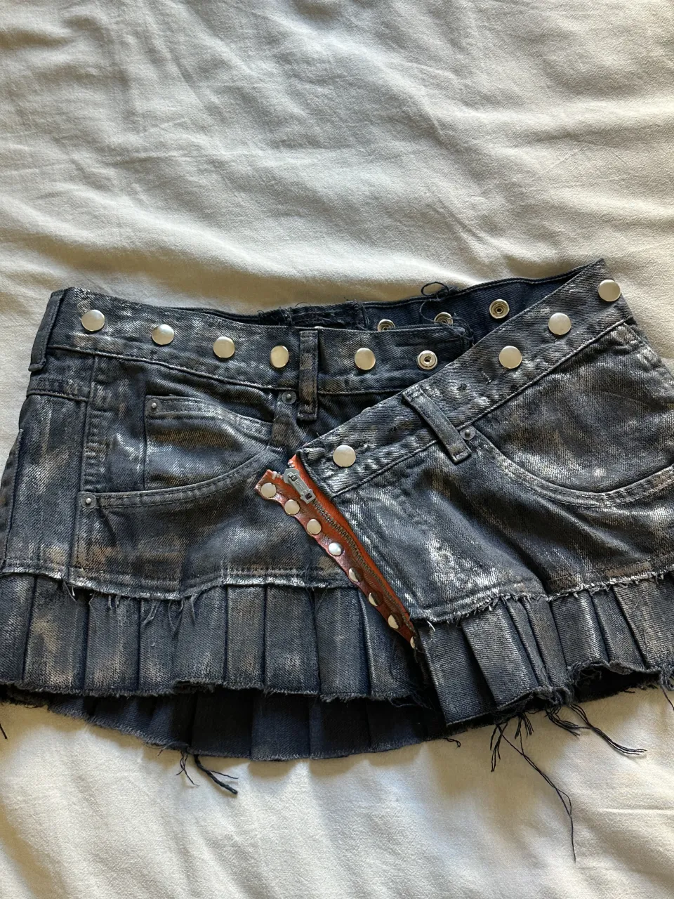 Pollera áurea de jean corta con detalles metálicos plateados y fruncidos. Posee cierre delantero y botones (uno fallado).