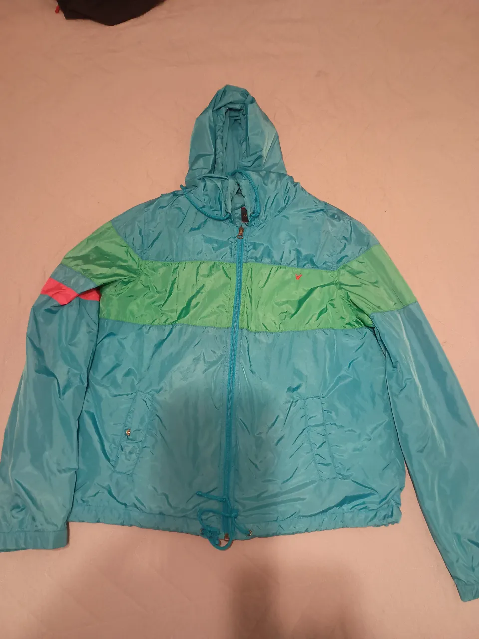 Campera deportiva con capucha, cierre al frente y dos bolsillos laterales. Diseño de bloques de color en turquesa, verde y detalles en rosa en las mangas.