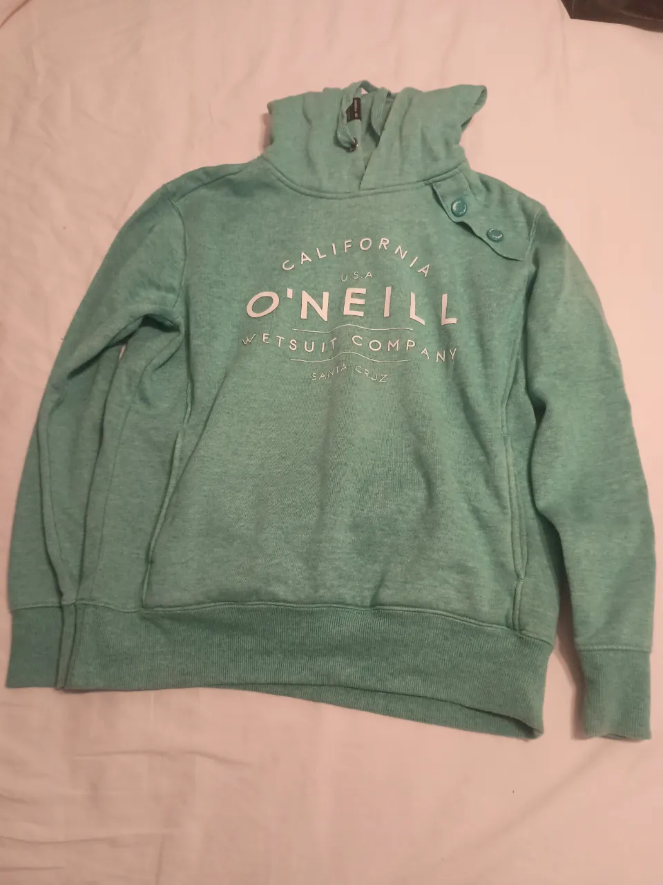 Buzo con capucha color verde agua, con estampa de la marca O'Neill en el frente. Tiene cordones en la capucha y un bolsillo canguro.