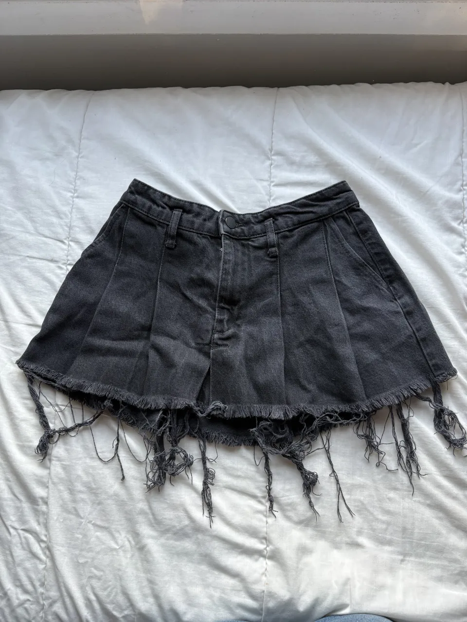 Pollera de jean negra con roturas y flecos en el ruedo. Posee pliegues en la parte delantera.