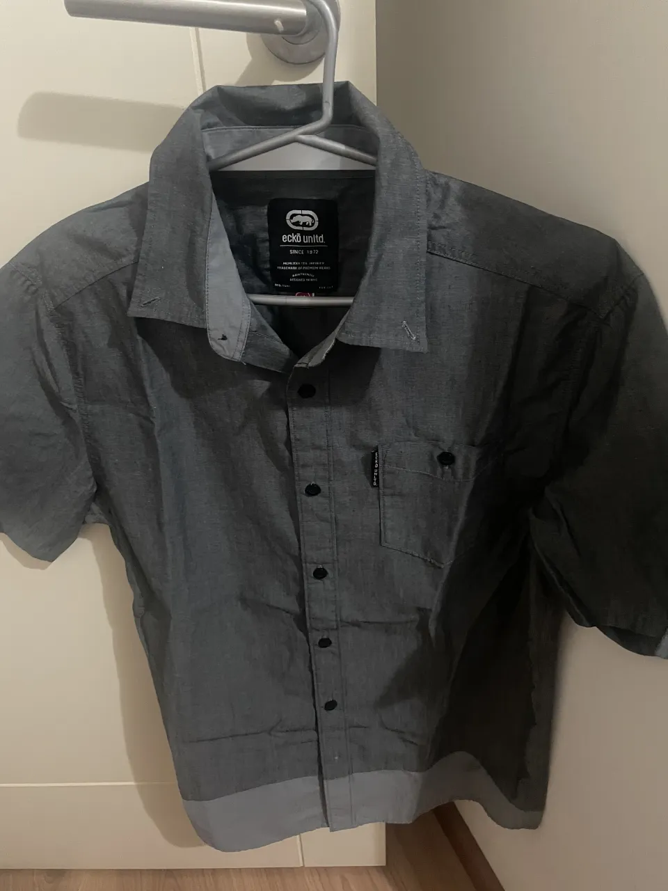 Camisa de vestir gris de manga corta, con botones al frente y un bolsillo en el pecho. Ideal para un look casual o semi-formal.