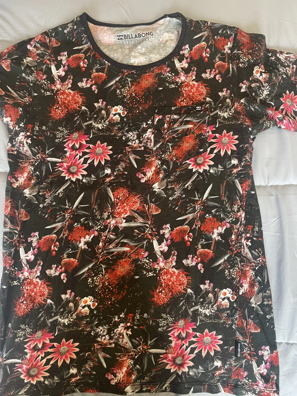 Remera Billabong Essentials para hombre, con estampado floral en tonos rojos y negros. Cuello redondo y bolsillo en el pecho. Talle M.