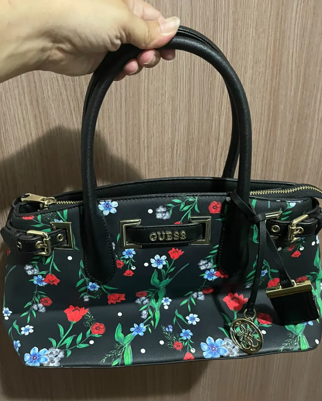 Cartera Guess negra con estampado floral. Diseño con manijas superiores y correa ajustable. Detalles dorados y cierre superior.