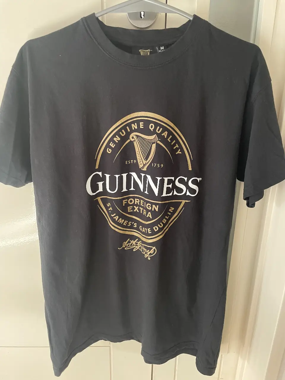 Remera negra de hombre con estampado del logo de Guinness en dorado y blanco. Cuello redondo y manga corta. Ideal para un look casual y cómodo.