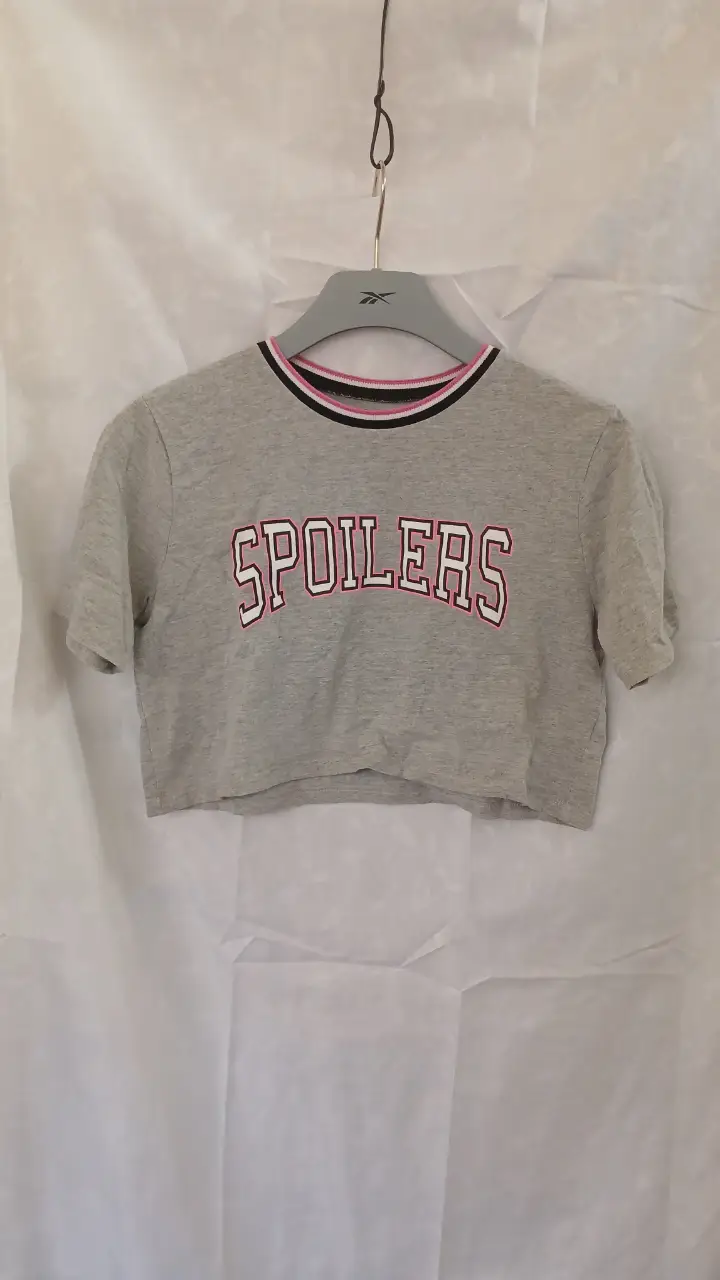 Remera crop gris con estampado "SPOILERS" en letras rojas y borde blanco. Cuello y mangas con detalles en rosa, blanco y negro. Ideal para un look casual y moderno. talle s