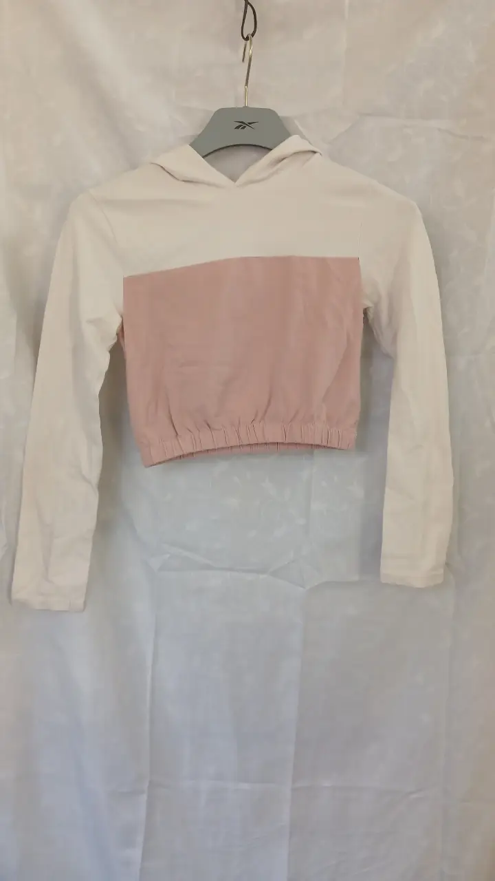 remera crop de niña, con capucha y mangas largas. Combina blanco y rosa en un diseño moderno y cómodo, ideal para un look casual y a la moda.