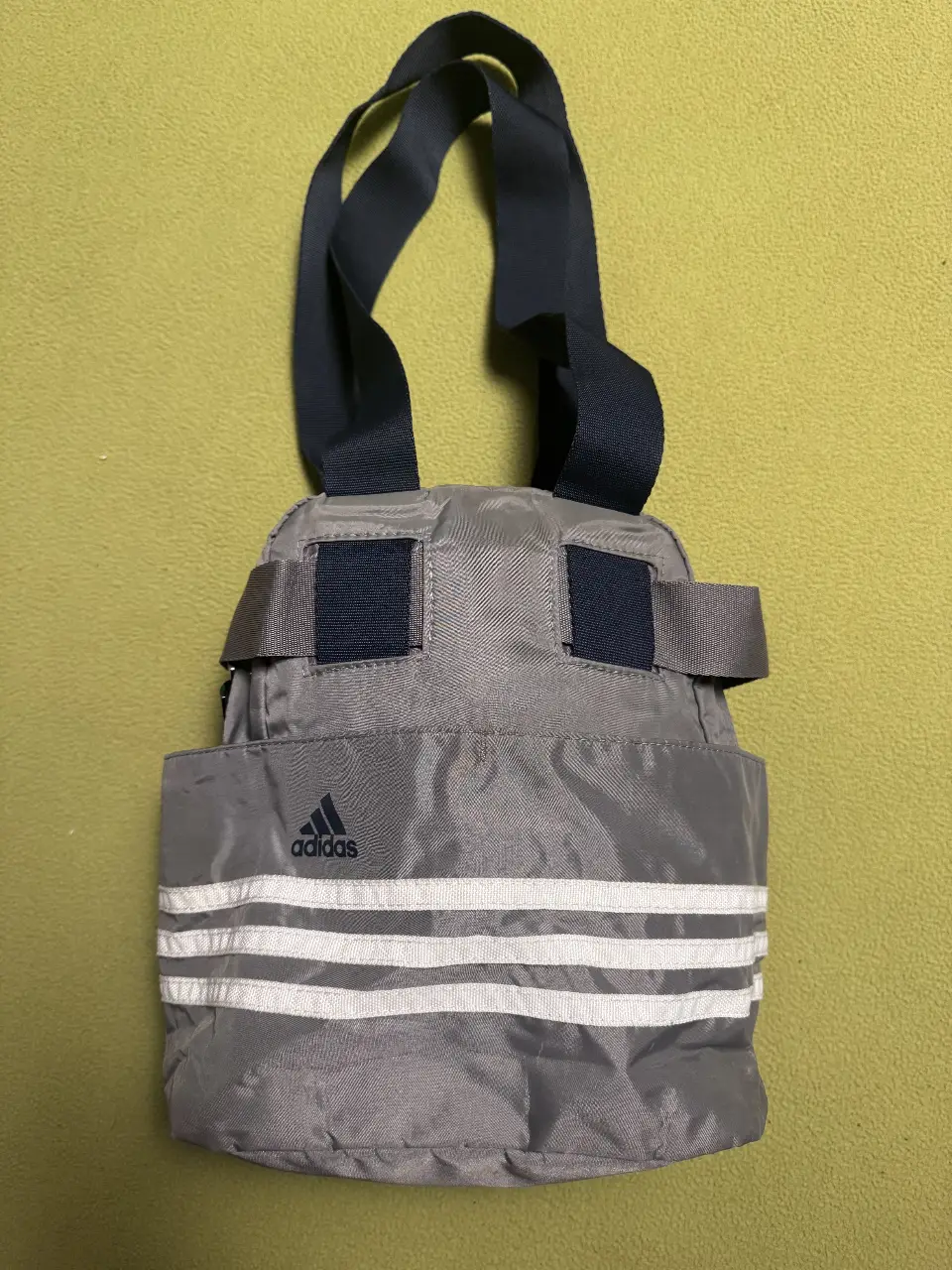 Bolso deportivo Adidas gris con detalles blancos. Ideal para llevar tus cosas al gimnasio o para actividades deportivas. Cuenta con tres rayas blancas características de la marca y un bolsillo frontal.