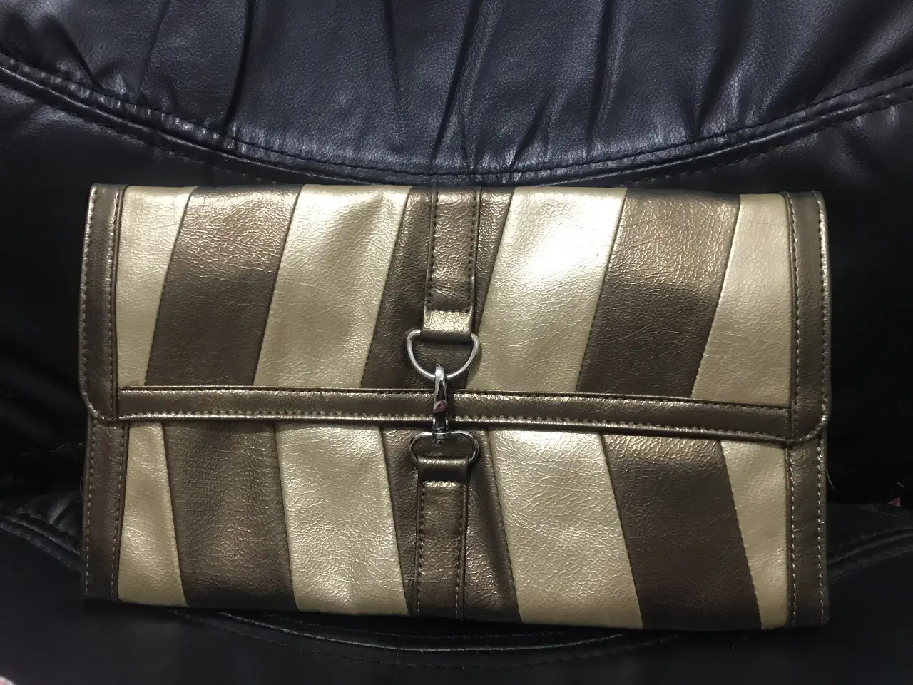 Cartera de mano con diseño a rayas diagonales en tonos dorados y marrones. Cuenta con un cierre y un detalle metálico en la solapa. Incluye una cadena para usar como bandolera.
