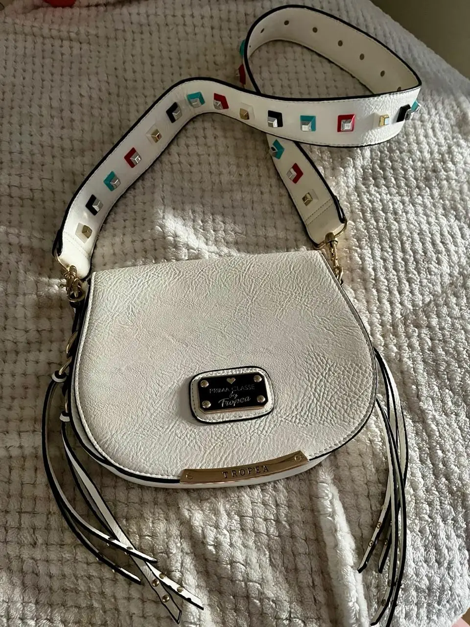 Cartera bandolera blanca con correa decorada. Diseño elegante con detalles dorados y flecos. Ideal para un look casual o de fiesta.