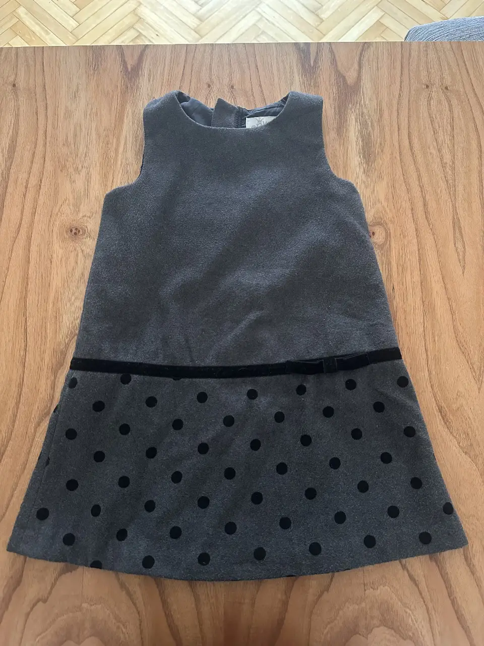 Vestido gris de niña con diseño de lunares negros en la falda. Corte recto, sin mangas y con detalle de cinta negra en la cintura. Ideal para ocasiones especiales.