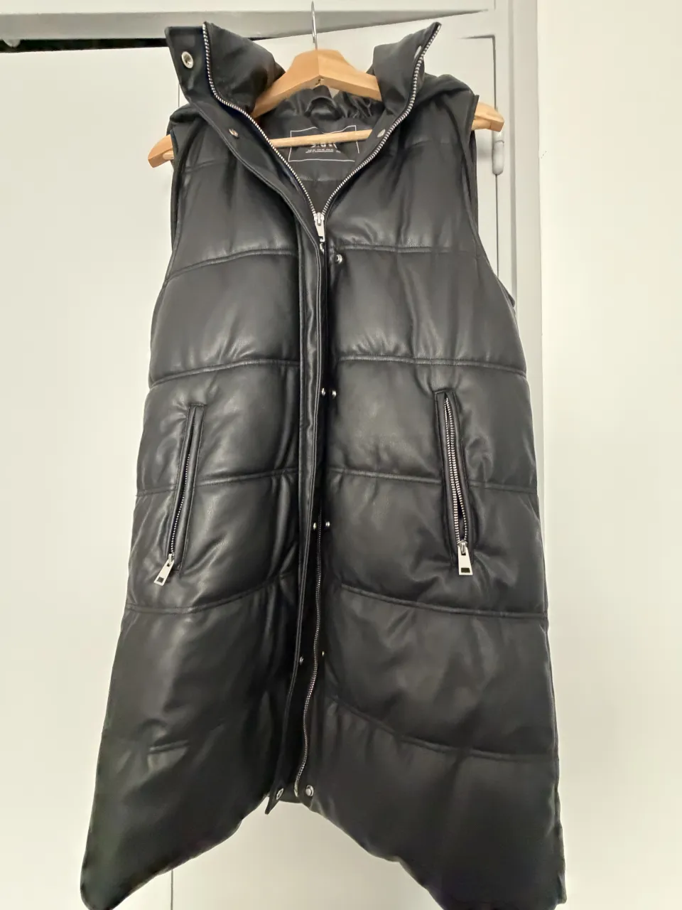 Chaleco acolchado negro con capucha, cierre al frente y bolsillos con cierre. Largo a la rodilla