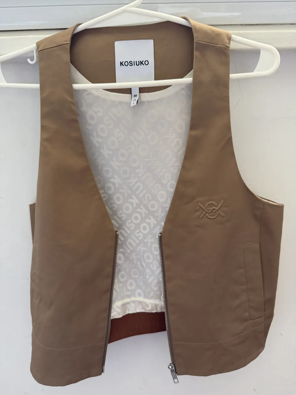 Chaleco de color marrón con cierre metálico al frente. Posee un bolsillo en cada lateral y un logo bordado en el frente del lado derecho. El interior está forrado con una tela blanca estampada con el logo de la marca.