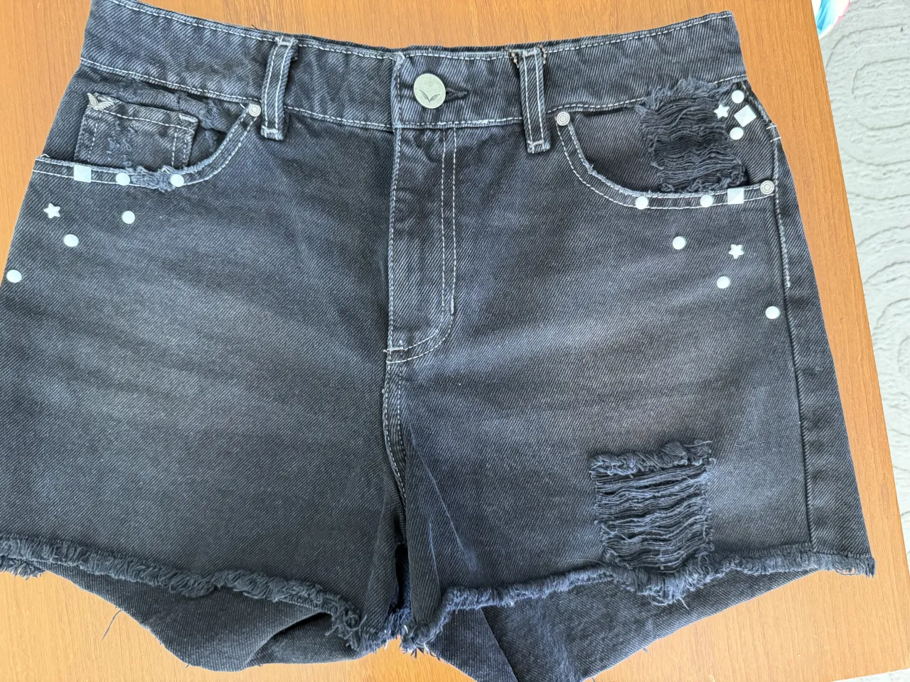 Shorts de jean negros con roturas y detalles de tachas con forma de  estrellas blancas pintadas. Tienen los bordes deshilachados.