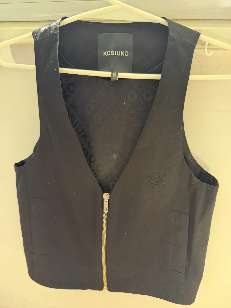 Chaleco negro con cierre metálico y logo bordado en el frente. El interior tiene un estampado repetido de la marca.