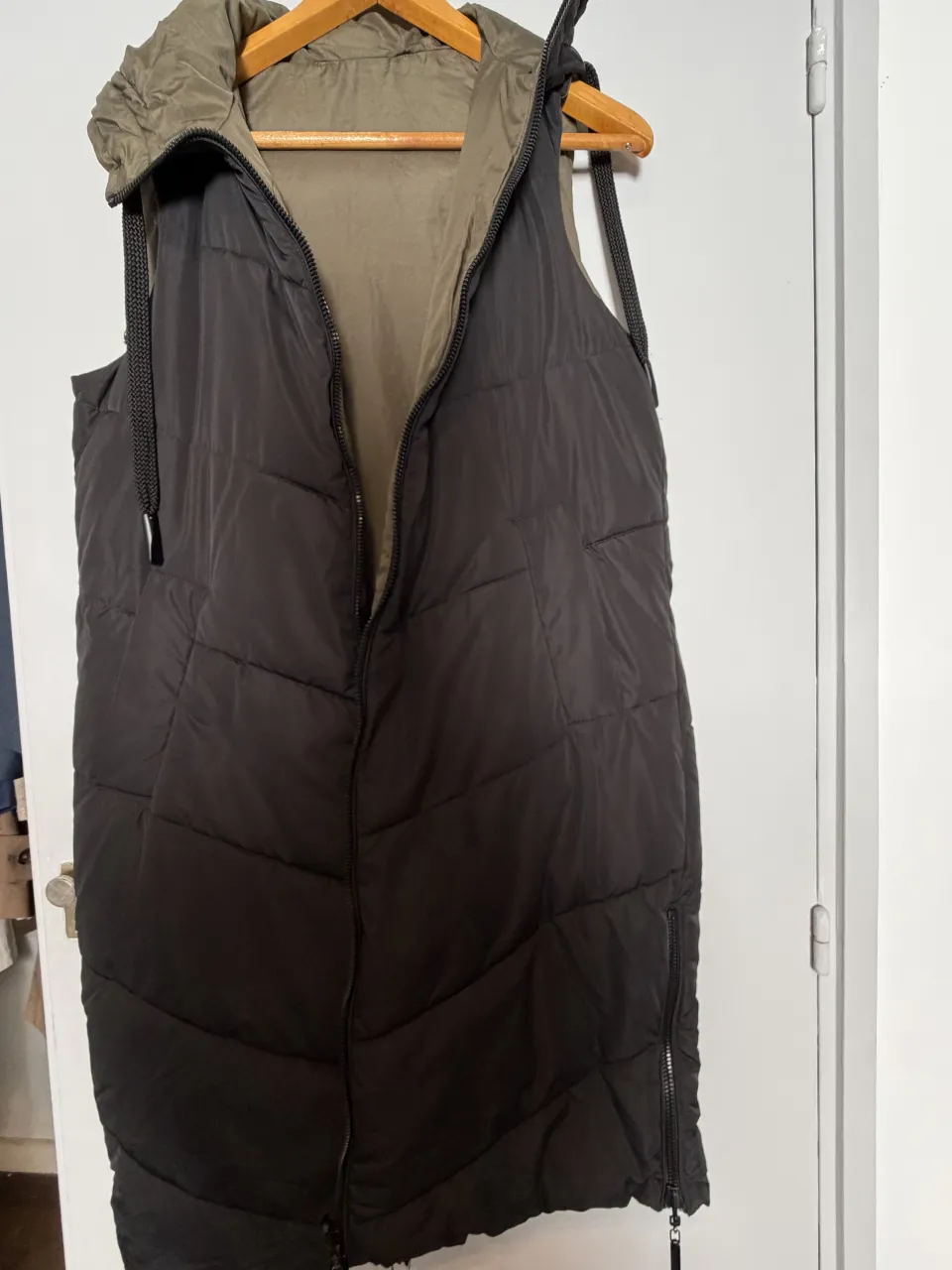 Chaleco acolchado largo con capucha. Tiene cierre al frente y bolsillos laterales con cierre. Es reversible, con un lado negro y otro verde oliva.