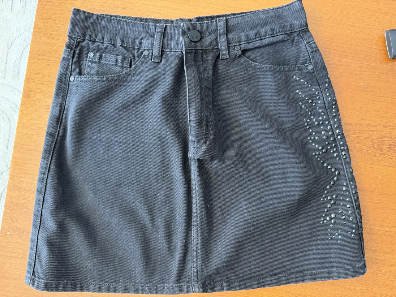 Pollera mini de jean negra con tachas plateadas en el lateral derecho. Tiene bolsillos delanteros y traseros.