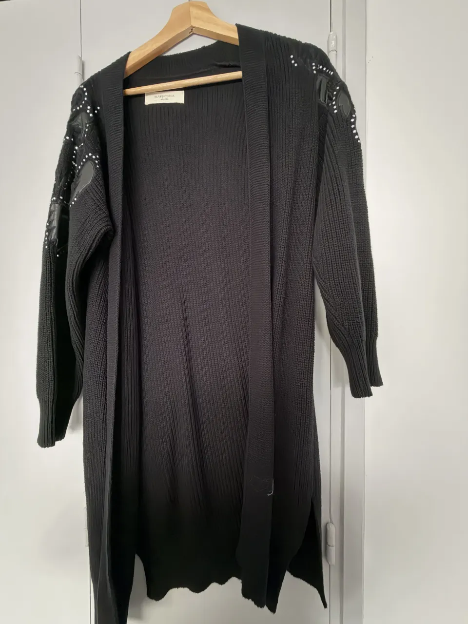 Cardigan largo de tejido negro con detalles de tachas plateadas y parches simil cuero en los hombros. Posee cuello redondo y mangas largas.