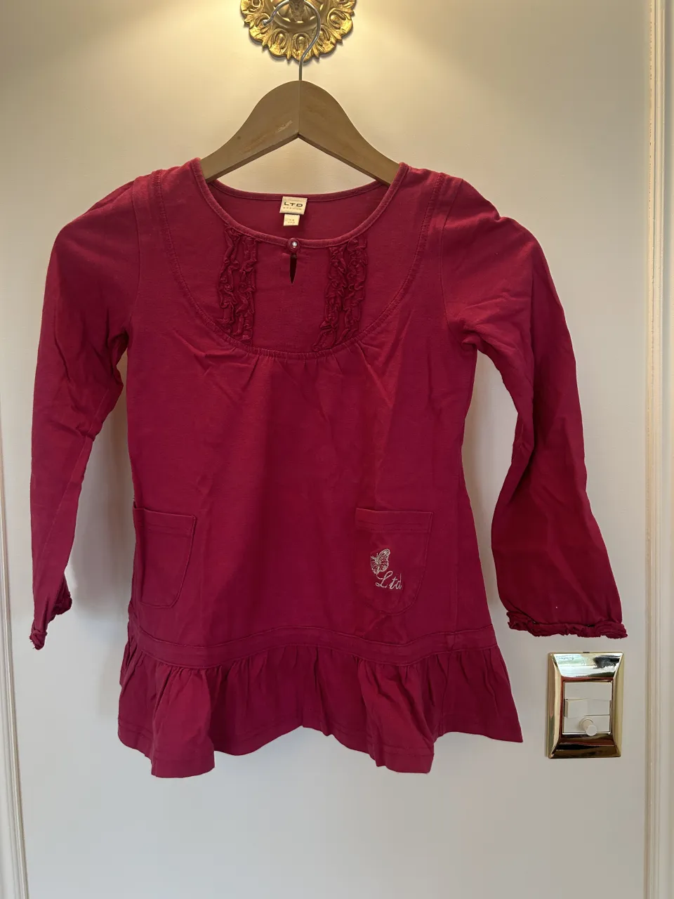 Remera manga larga de color fucsia con cuello redondo y detalle de volados en el frente. Posee un bolsillo aplicado en el frente izquierdo con bordado de mariposa y la inscripción "LTD". El ruedo es con volados.