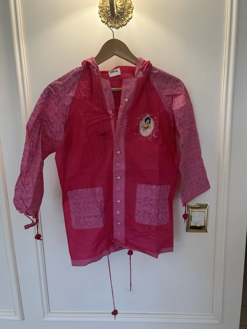 Impermeable rosa con mangas estampadas con flores. Posee capucha, bolsillos delanteros y estampa de princesas Disney en el frente y la espalda. Tiene un pequeño desperfecto en el puño izquierdo como indica la última foto 