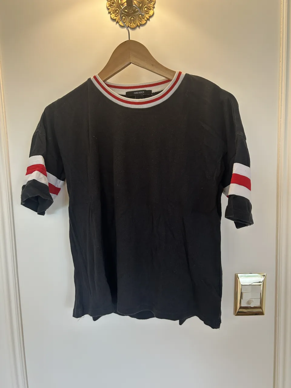 Remera negra con cuello redondo y mangas cortas. Presenta detalles de franjas rojas y blancas en el cuello y en los puños de las mangas. En la espalda, lleva estampado "NEW YORK" y el número "76" en blanco.