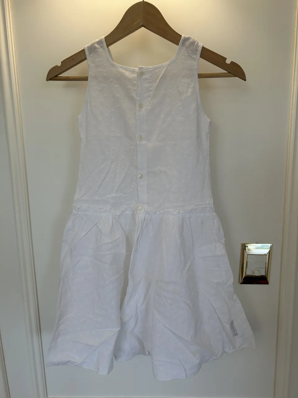 Vestido blanco sin mangas con botones en la espalda y detalle de volado en la cintura. Posee pequeñas texturas en relieve. Tiene Manchitas amarillas como se adjunta en la última foto 