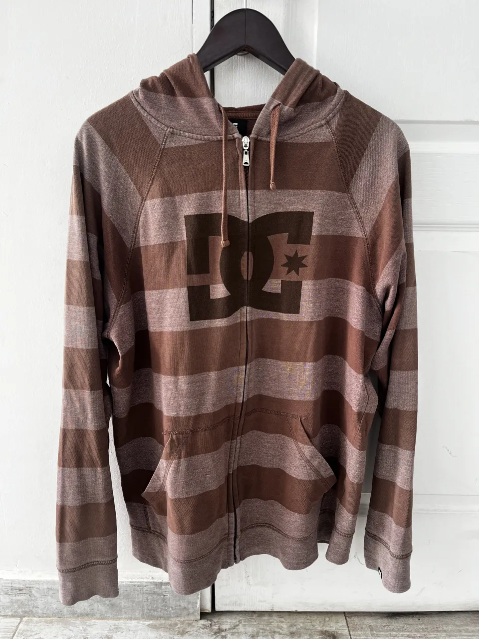 Buzo con capucha y cierre completo, de algodón a rayas marrones y beige. Tiene el logo de DC Comics estampado en el frente y bolsillos tipo canguro. Ideal para un look casual y urbano.