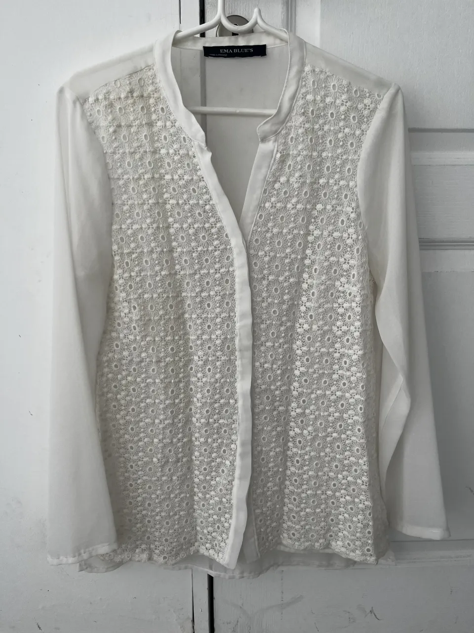 Blusa blanca de manga larga con detalles de encaje floral en el frente. Cuello mao y botones al tono. Transparente, ideal para un look elegante y delicado.