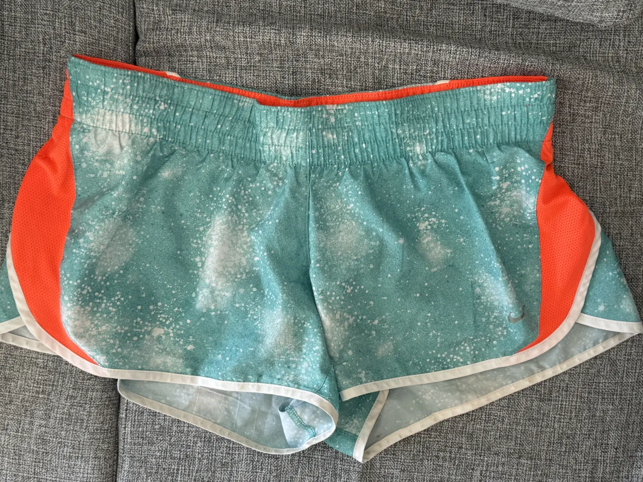 Shorts deportivos Nike Dri-Fit con un diseño vibrante y moderno. El color principal es un verde claro con salpicaduras blancas, combinado con detalles laterales en naranja brillante y ribetes blancos. Ideales para entrenar o para un look casual y deportivo. es talle L pero puede ir para un talle M.