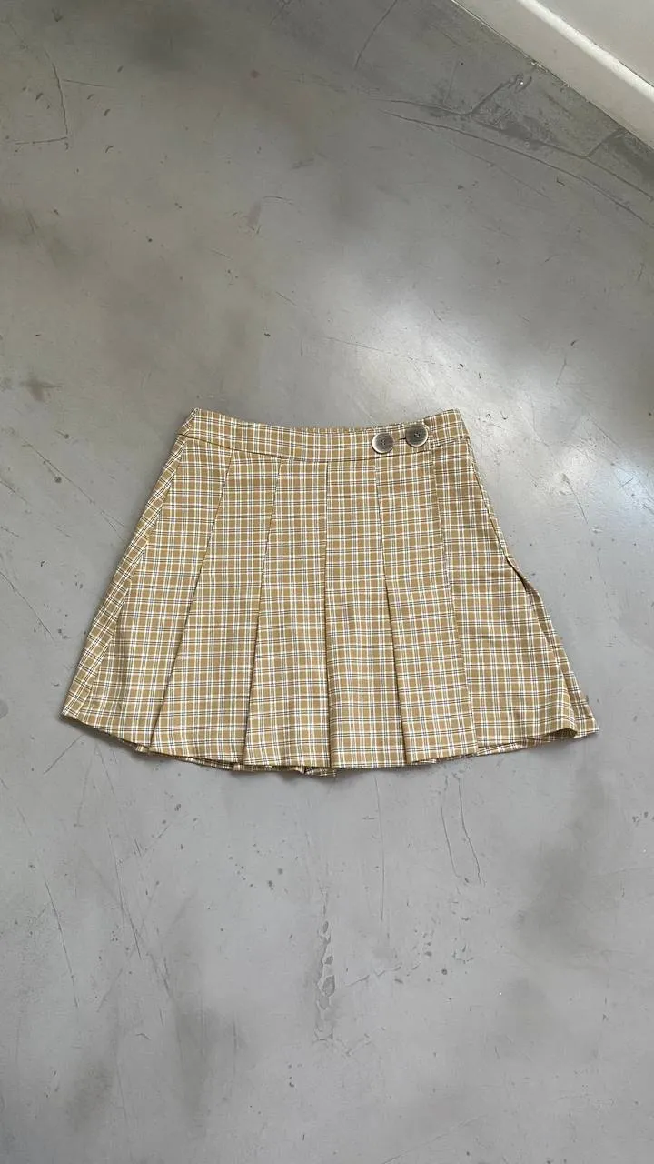 Pollera corta tableada [cuadros escoceses en tonos mostazas y blanco]. Posee pliegues y dos botones para abrochar en la cintura. Medidas: cintura 35cm - cadera 44cm - largo 39cm. 