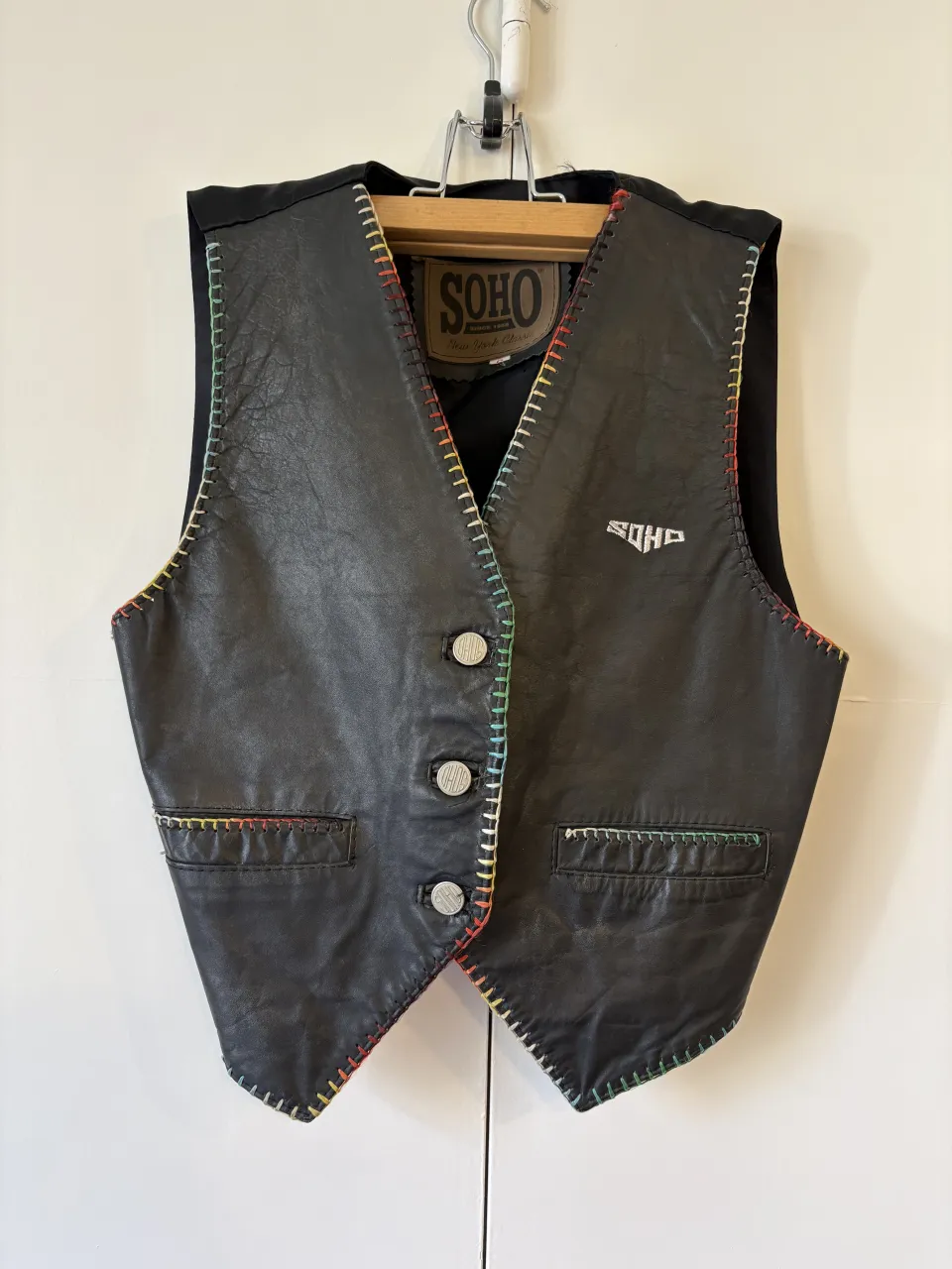 Chaleco de cuero sastrero negro con costuras de colores en los bordes y bolsillos. Cuero bien rigido, vintage. Posee tres botones frontales y un logo bordado en el frente.