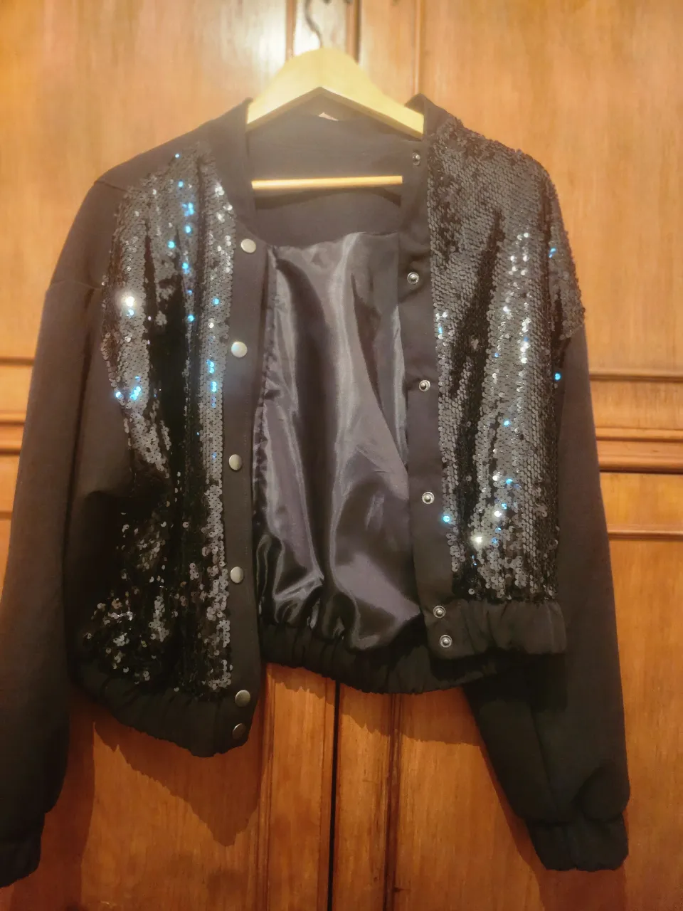 Campera bomber negra con lentejuelas brillantes en la parte delantera y mangas. Cierre de broches metálicos y puños y cintura elastizados.