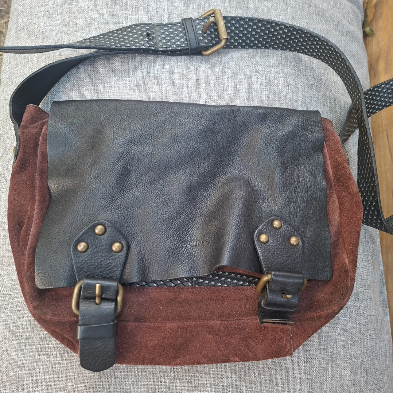 Cartera de cuero y gamuza marrón con solapa de cuero negra. Posee hebillas y tachas metálicas. Correa regulable.