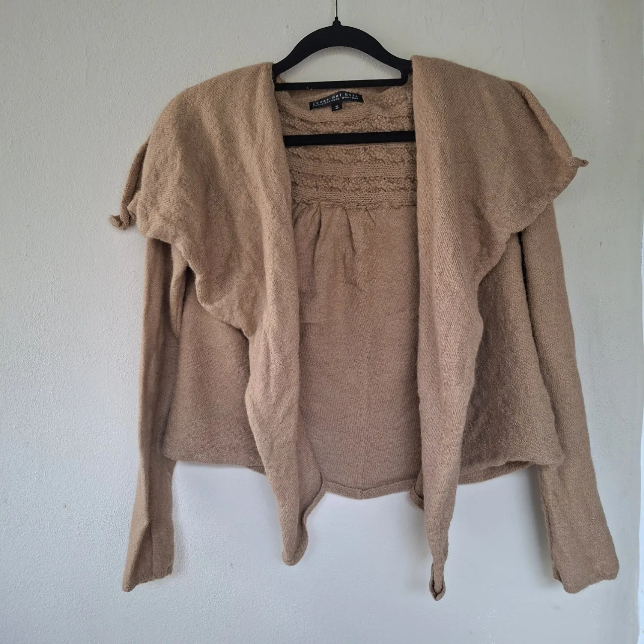 Cardigan de lana color beige con mangas largas y cuello solapa. Posee detalles de tejido trenzado en la espalda y en la parte superior del frente. Se abre al frente.