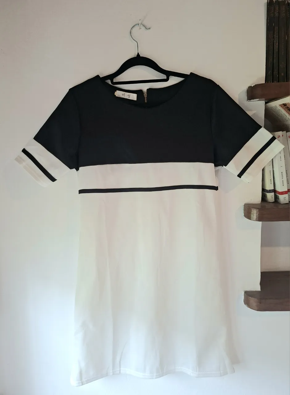Vestido corto de mangas cortas, con cuello redondo y cierre en la espalda. Presenta un diseño de bloques de color, con la parte superior y las mangas en negro, y la parte inferior en blanco. Las mangas tienen dos finas líneas horizontales negras en los bordes. Una línea horizontal negra divide la parte superior de la inferior.