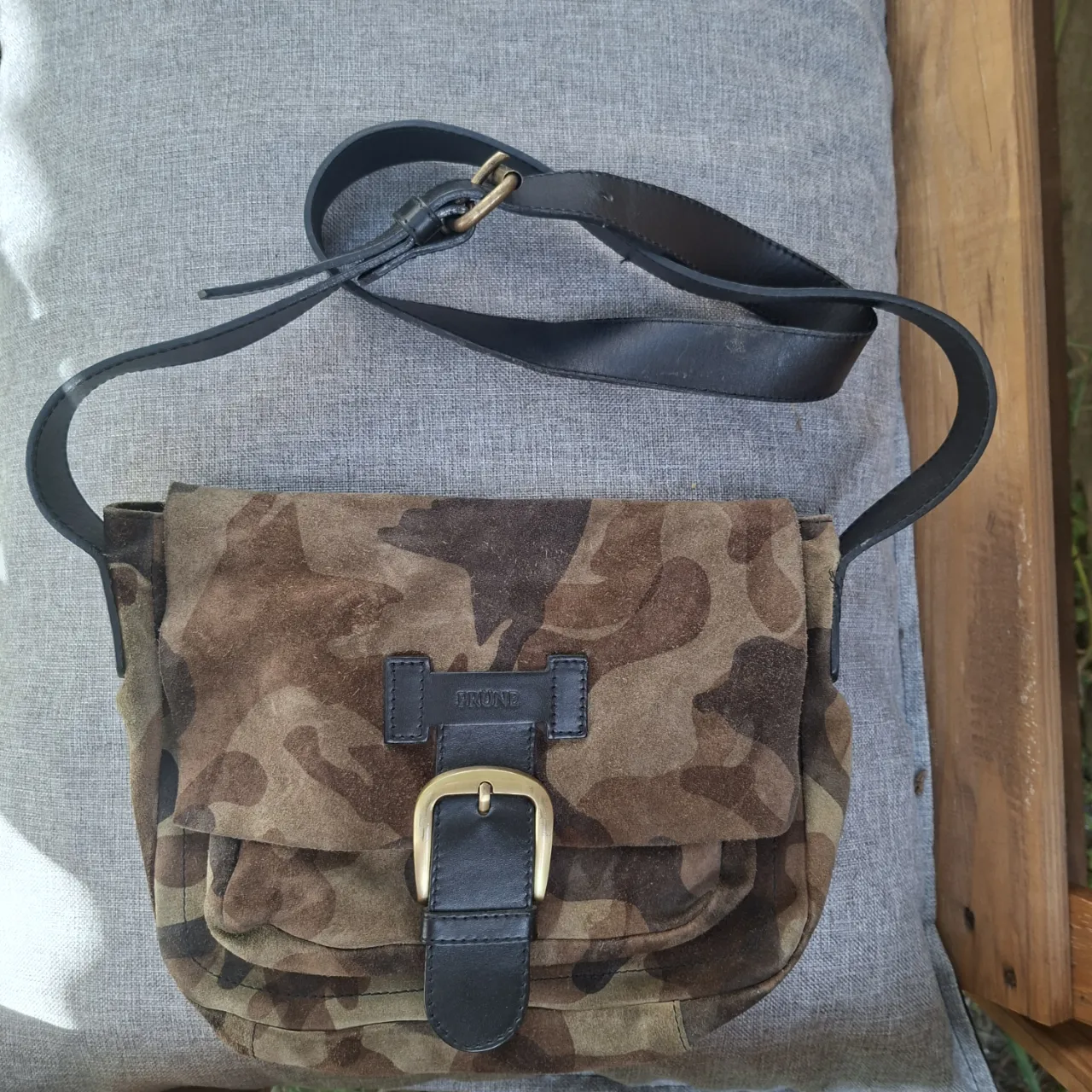 Cartera bandolera de cuero y gamuza con estampa camuflada. Tiene hebilla dorada y detalles en cuero negro. Ideal para un look casual y moderno.